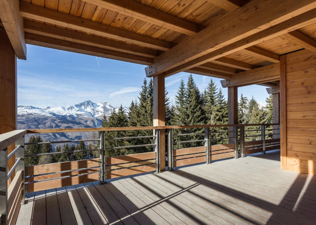 Grand balcon en bois de chalet avec vue imprenable sur des montagnes enneigées et une forêt de sapins sous un ciel bleu éclatant.