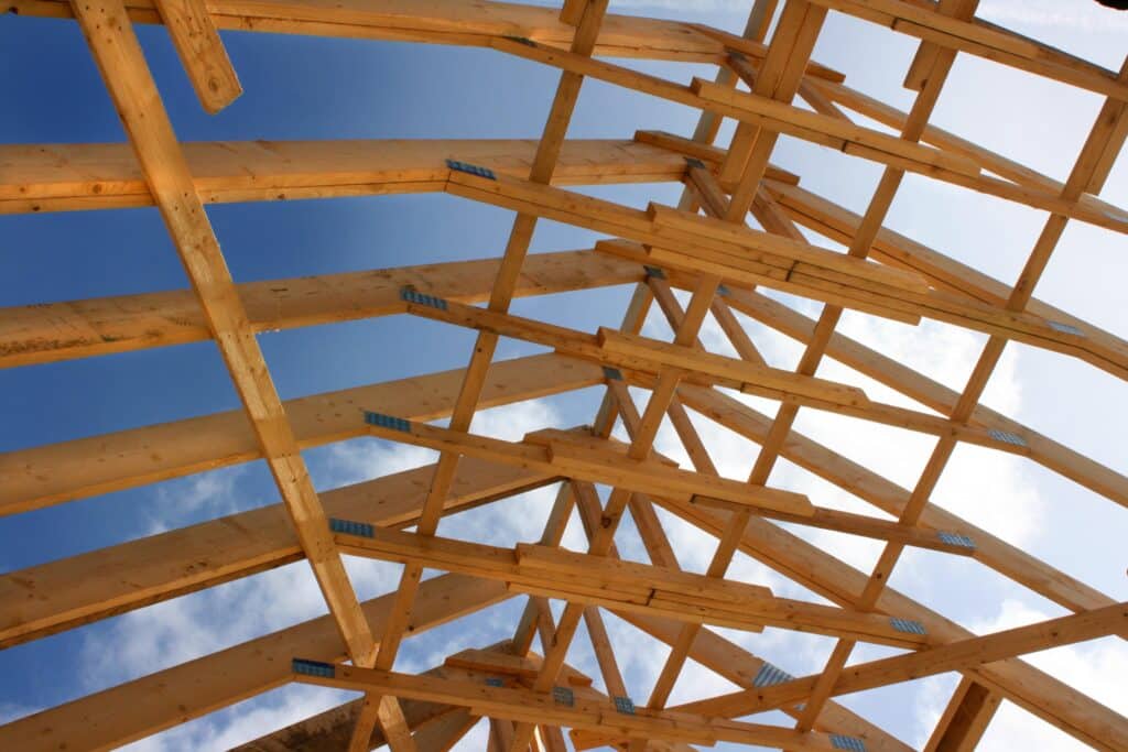 Charpente de toit en bois avec des fermes complexes sous un ciel bleu clair et quelques nuages, vue du bas sur un chantier.