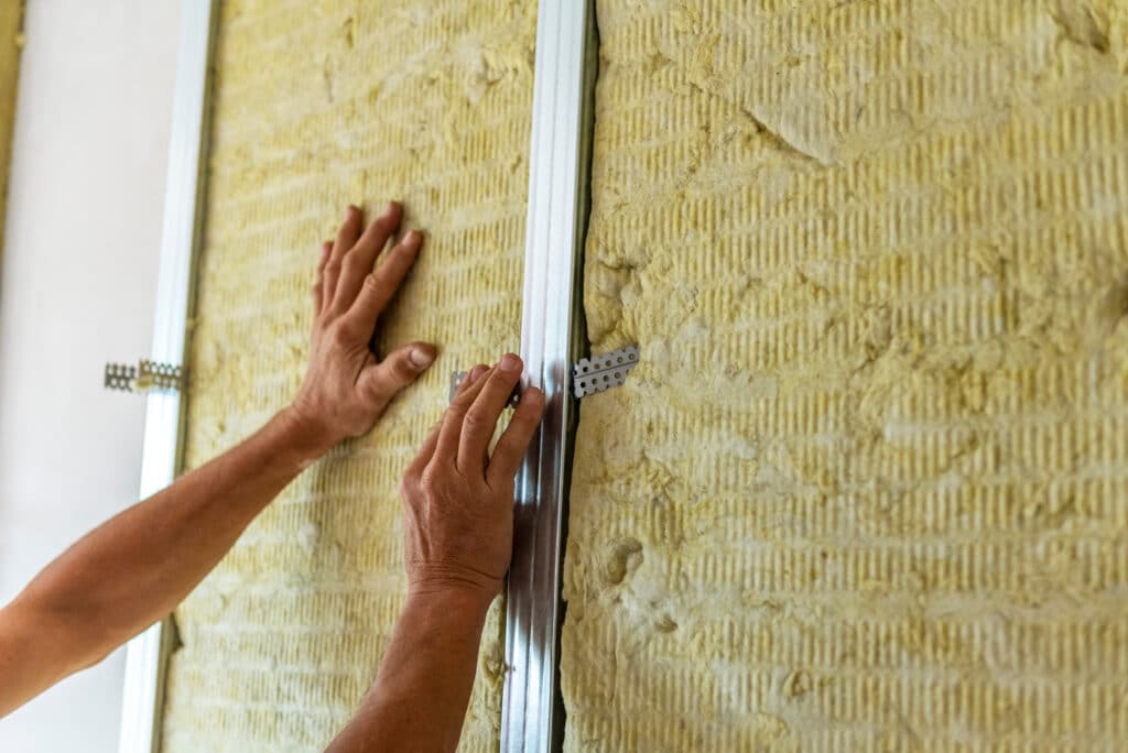 Des mains installent de la laine de roche sur un mur, fixée par des profils métalliques pour l'isolation thermique.